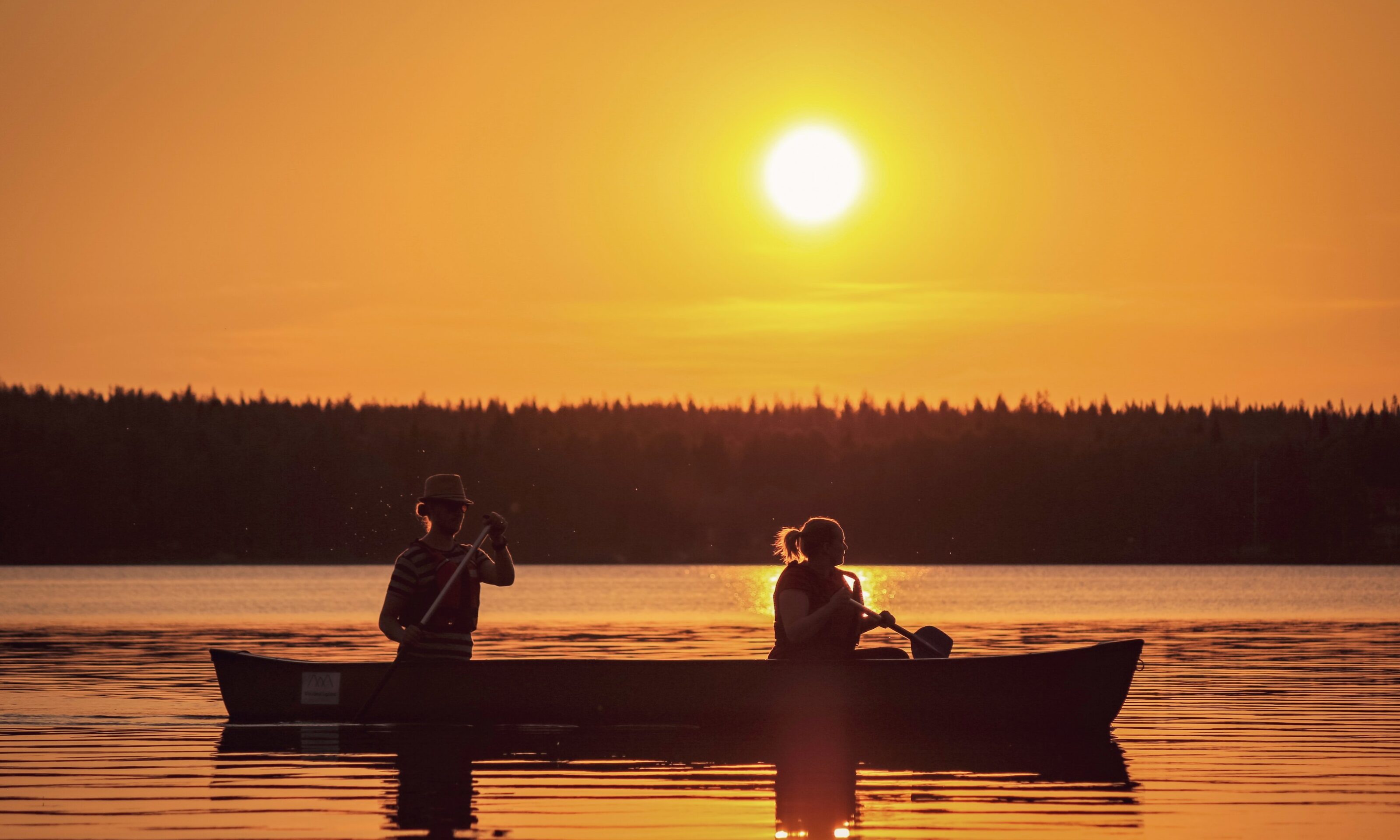 Two people in a canoe on a calm lake at sunset, with the golden sun low on the horizon, reflecting on the water. The scene evokes a tranquil, Arctic summer moment.