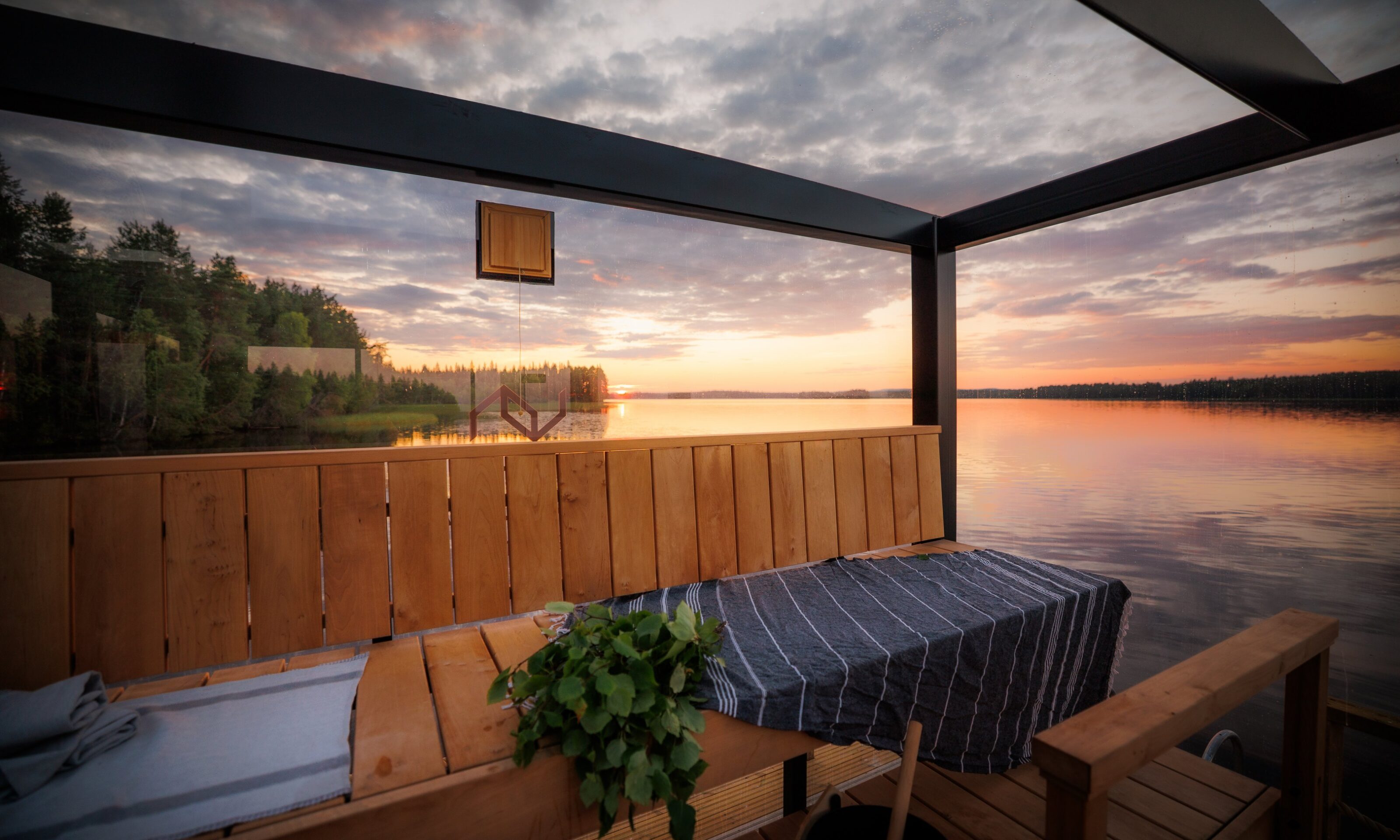A glass-walled sauna opens onto a lakeside view at sunset. Wooden benches, a sauna whisk and the softly lit sauna interior create a calm, warm atmosphere.