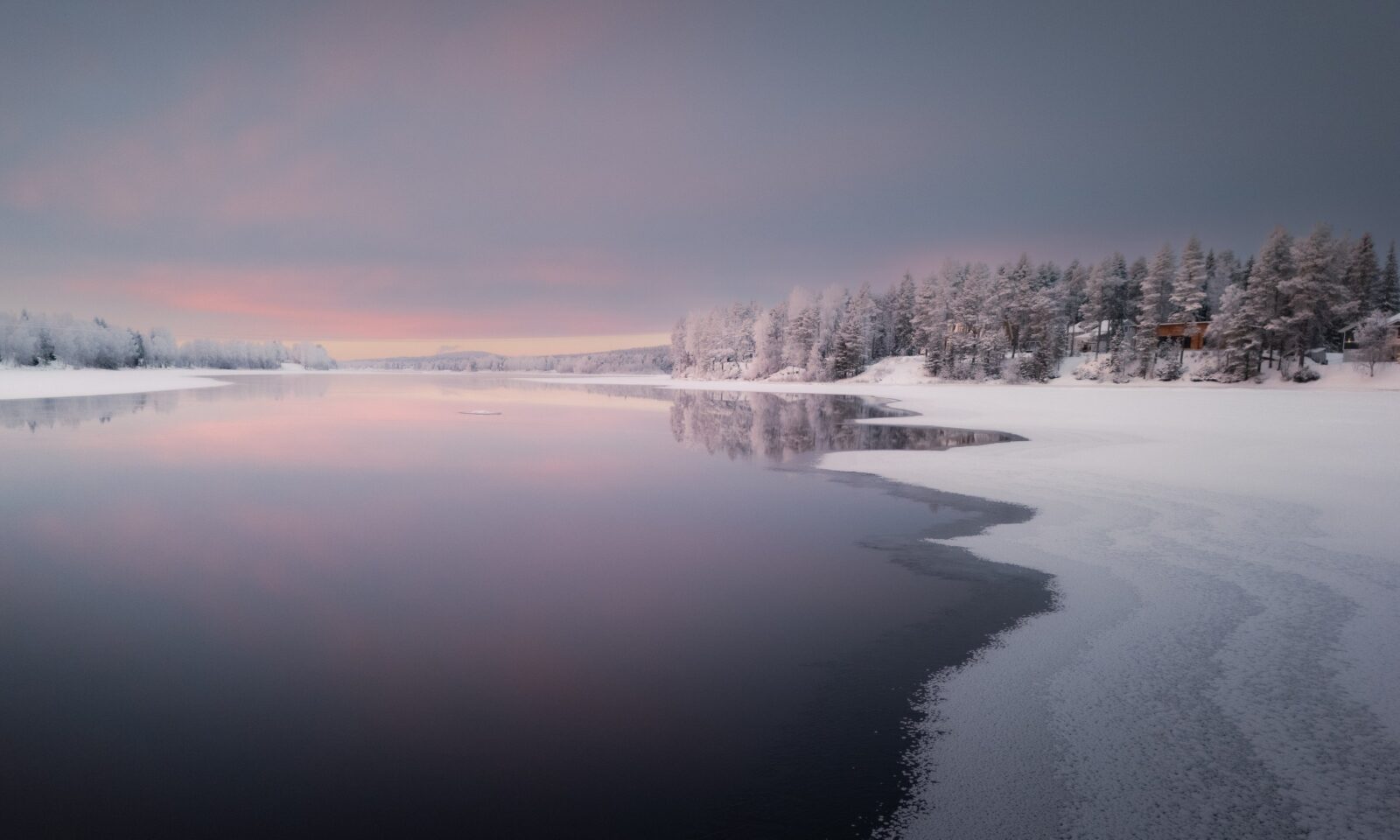 Snow-covered shoreline and calm winter water under a soft pink sky at Aino Private Island in Rovaniemi, Finnish Lapland.